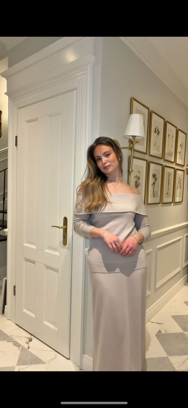 Woman in a gray off-shoulder dress standing in a decorated room with white walls and framed pictures.